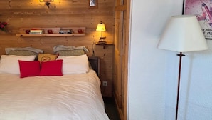 Un dormitorio acogedor con una pared de madera, una cama con ropa de cama blanca y almohadas rojas, una mesita de noche con una lámpara y un estante con libros y objetos decorativos.