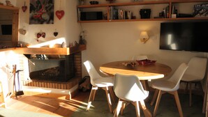 Un salón acogedor con chimenea, una mesa redonda de madera rodeada de sillas y una estantería llena de libros y objetos decorativos.