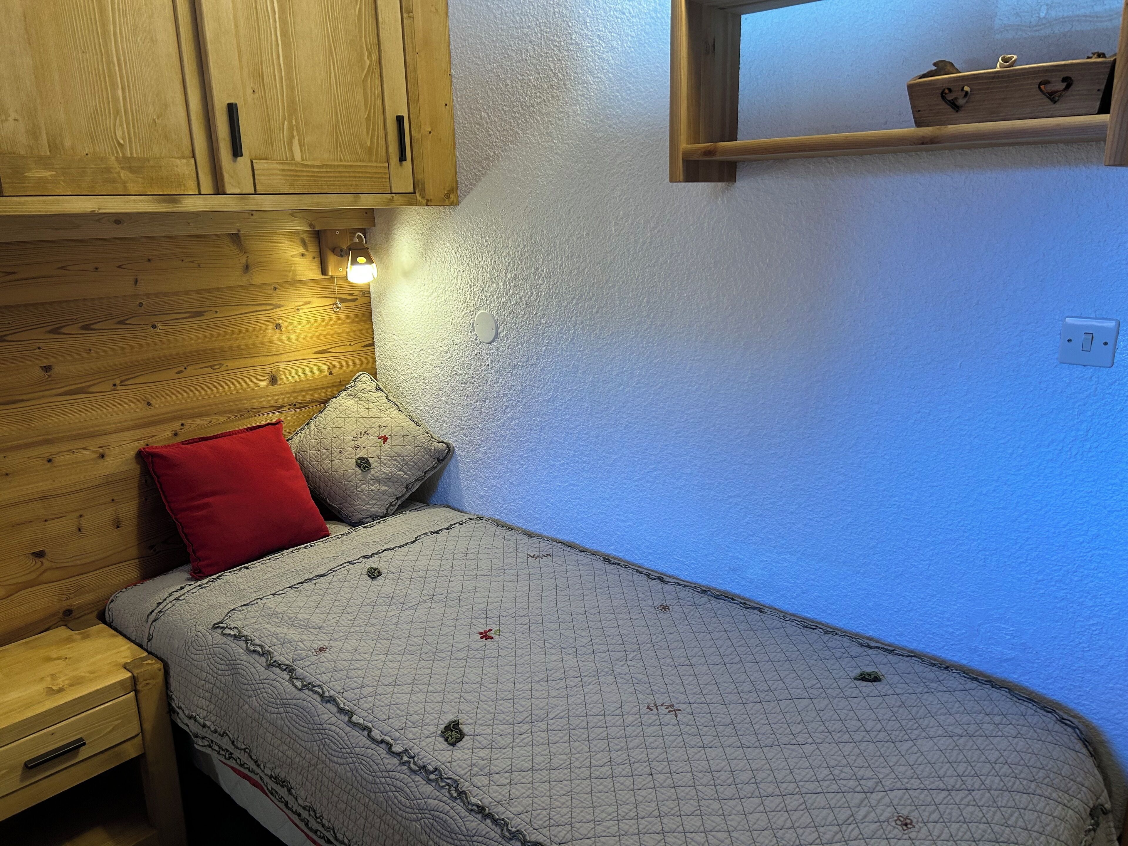 Ein kleines, gemütliches Schlafzimmer mit einem Holzkopfteil, einem Einzelbett mit gesteppter Bettdecke, einem roten Kopfkissen und einem kleinen Regal über dem Bett.