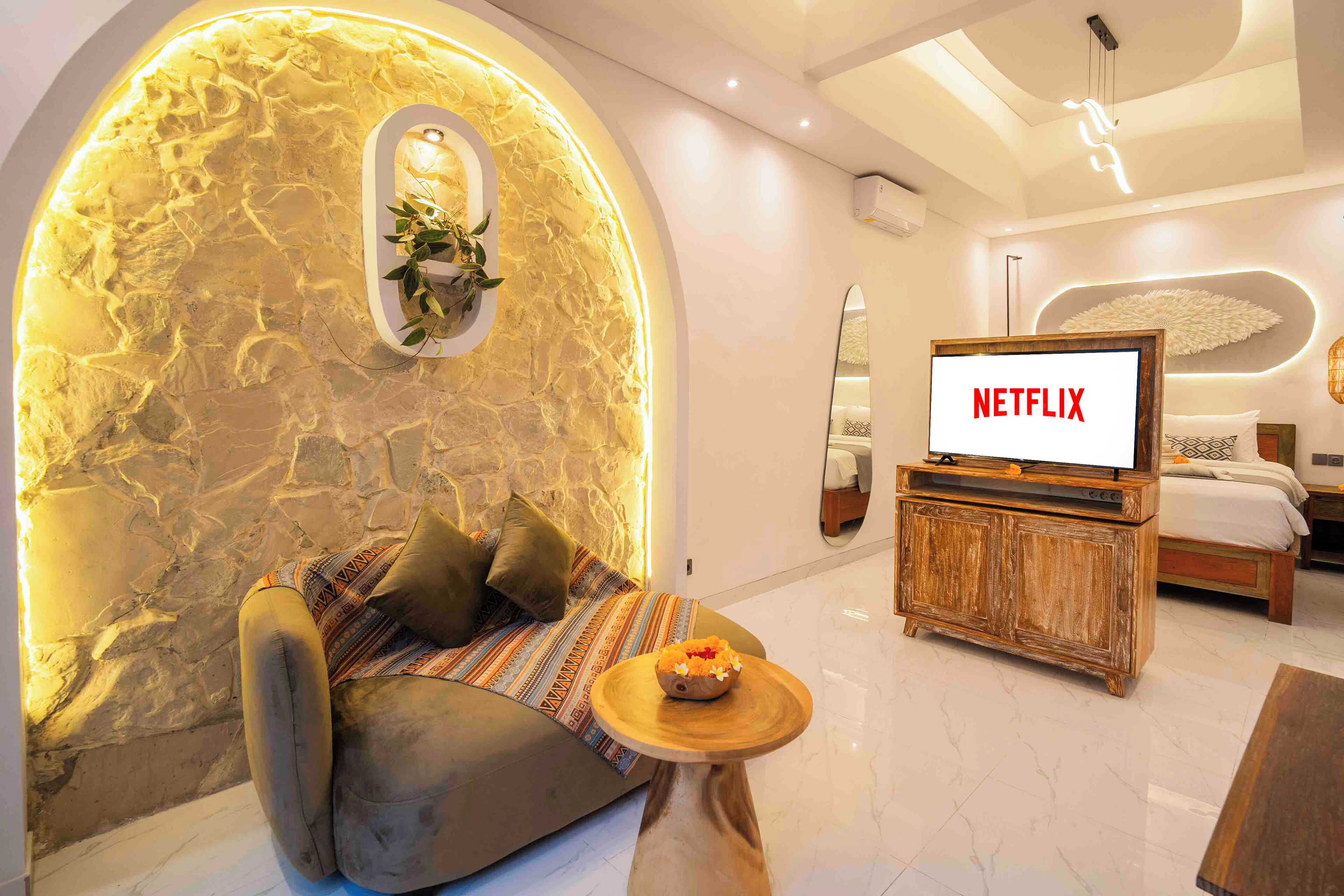 Une chambre d’hôtel moderne avec un mur en pierre, un coin salon confortable, une table basse en bois, un lit et un téléviseur affichant le logo de Netflix.