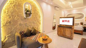 Une chambre d’hôtel moderne avec un mur en pierre, un coin salon confortable, une table basse en bois, un lit et un téléviseur affichant le logo de Netflix.