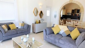 A modern living room with two grey sofas, a glass coffee table, and a kitchen visible through an archway.