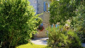 Un bâtiment en pierre avec un chemin de gravier menant à une porte, entouré d’une végétation luxuriante et d’un petit jardin.
