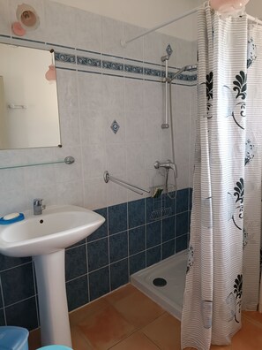 Une salle de bain avec un lavabo blanc, une étagère en verre, une douche avec un rideau blanc et des murs recouverts de carreaux bleus et blancs.