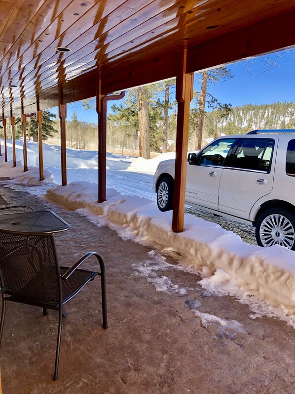 Un espacio al aire libre cubierto con techo de madera, sillas de metal y un SUV blanco estacionado cerca.