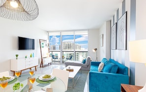 A modern living room with a blue sofa, a dining table with plates and glasses, and a large window offering a city view. - Vibrant Brickell Suite | Walk Everywhere (Miami)