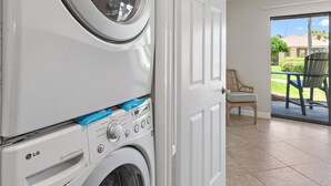 Une buanderie équipée d’un lave-linge et d’un sèche-linge à chargement frontal, une porte donnant sur un balcon avec une chaise et une table, et une vue sur un quartier résidentiel.