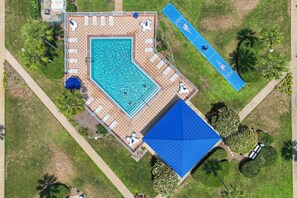 Vista aérea de una zona de piscina con un edificio de techo azul, tumbonas y una cancha de tenis.