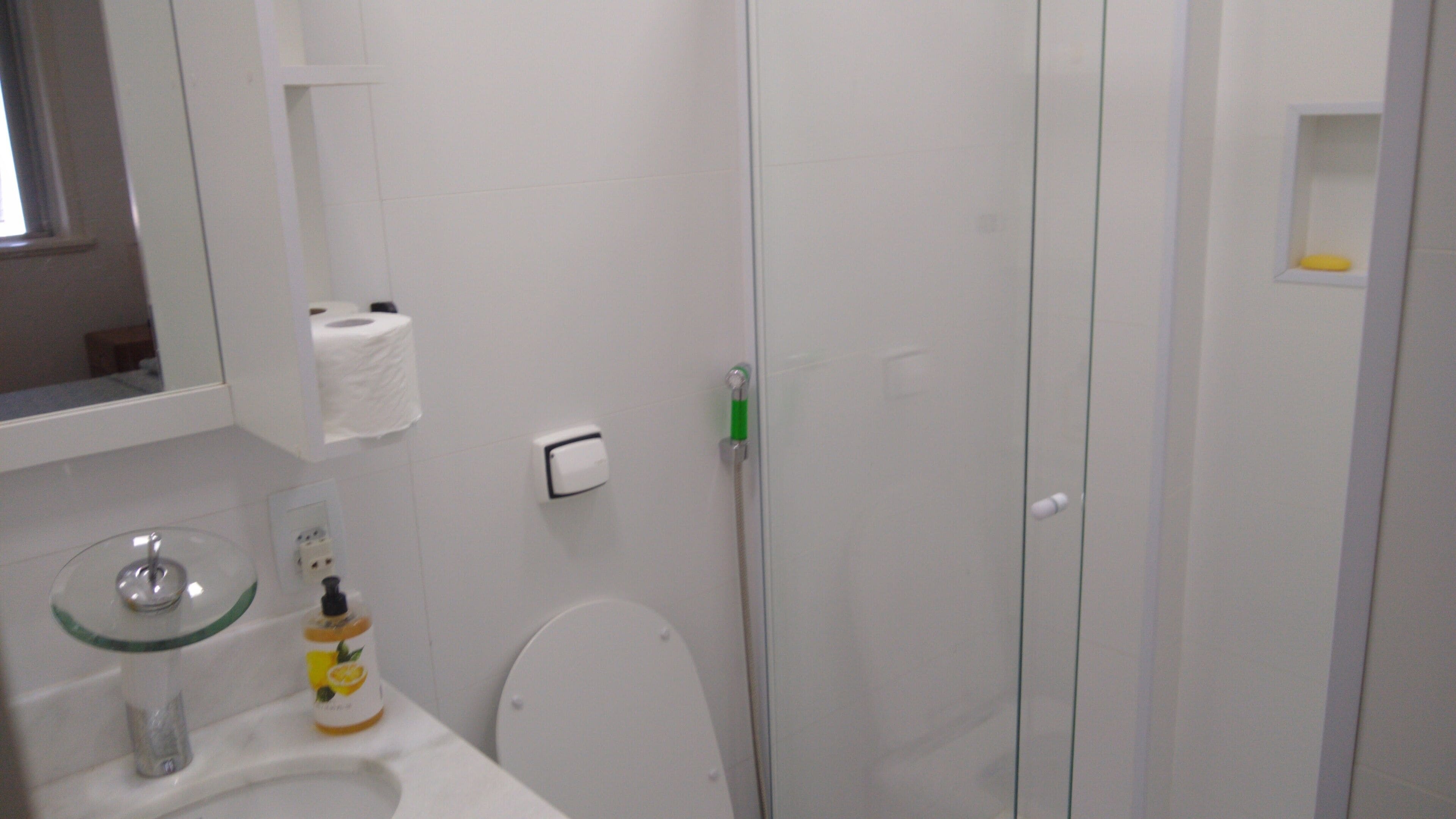 Un baño con inodoro, lavamanos de vidrio, espejo, dispensador de toallas y ducha con puerta de vidrio.