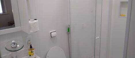 Un baño con inodoro, lavamanos de vidrio, espejo, dispensador de toallas y ducha con puerta de vidrio.
