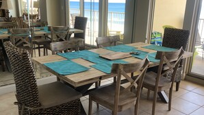 Un espace repas avec des tables et des chaises en bois, des sets de table de couleur turquoise et une vue sur l’océan grâce à de grandes fenêtres.