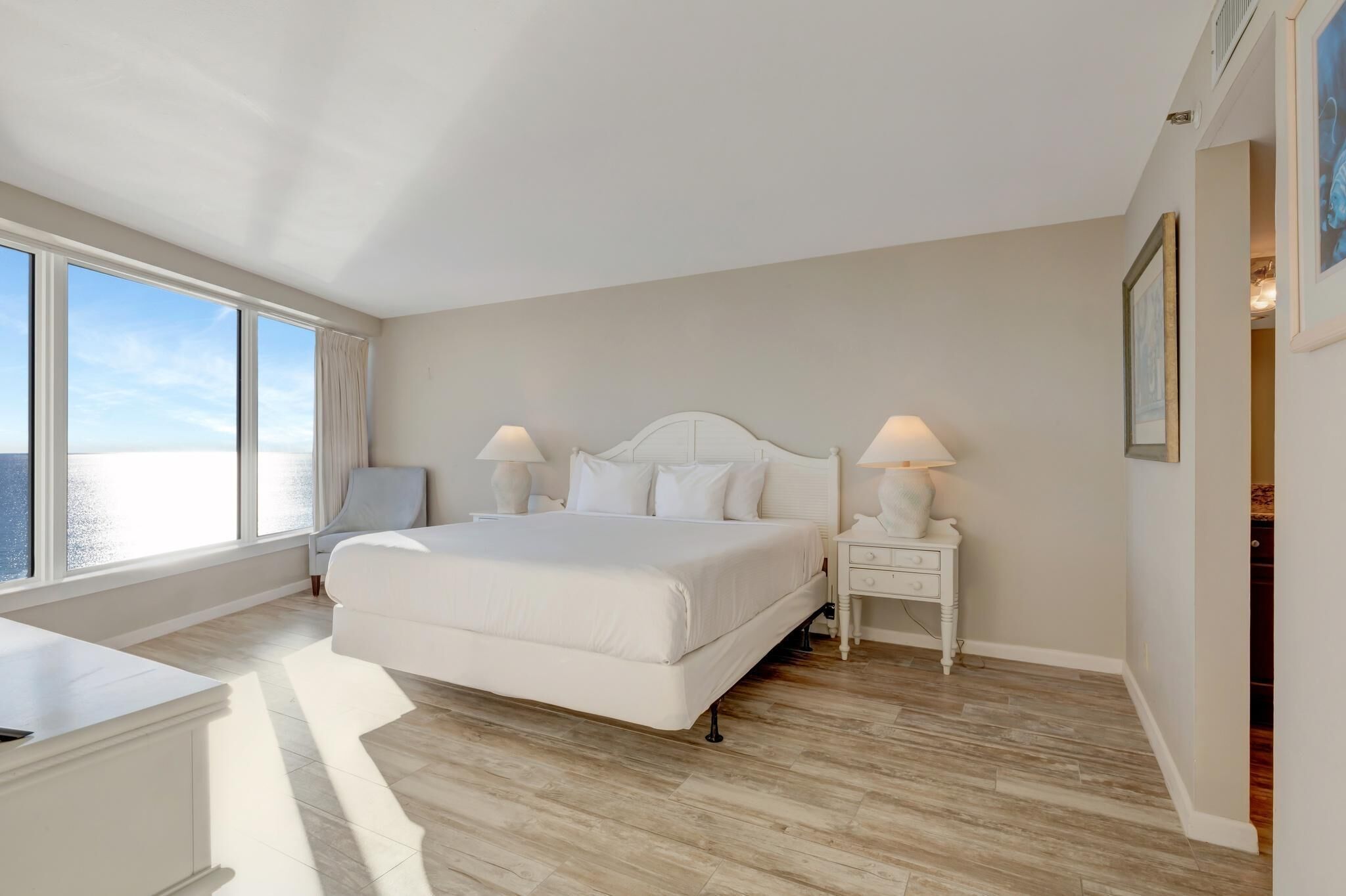 A spacious bedroom with a large bed, two bedside tables with lamps, a nightstand, and a view of the sea through large windows.