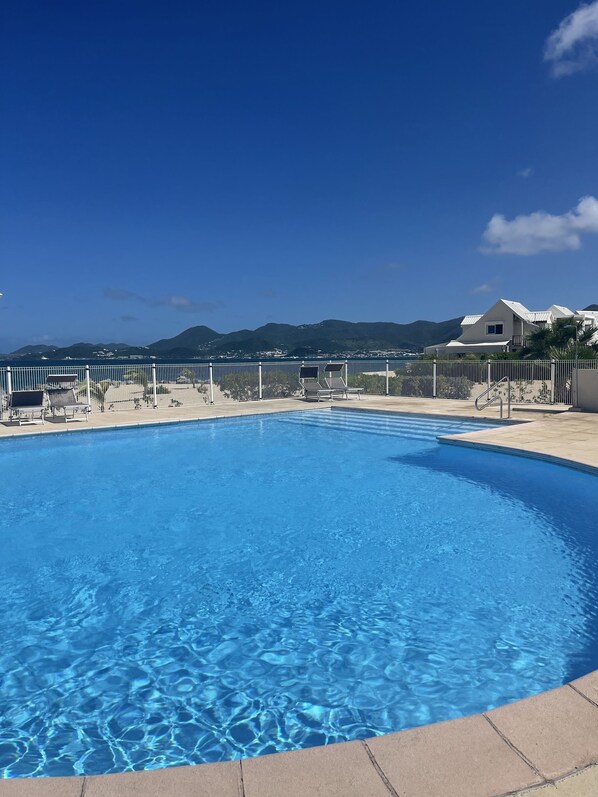Una piscina azul cristalina con una valla metálica, rodeada por una terraza pavimentada y sillones de descanso. Al fondo, se observan casas y un paisaje montañoso bajo un cielo despejado.