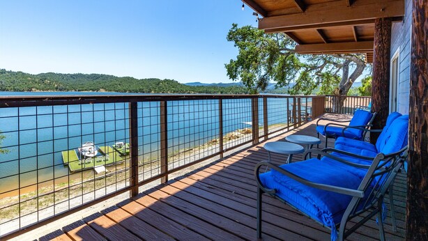 Une terrasse en bois avec des chaises longues bleues et une table, donnant sur un lac, avec un quai et de la verdure en arrière-plan.