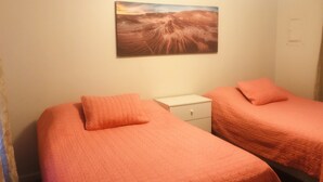 Ein Zimmer mit zwei Betten, jeweils mit einer orangefarbenen Bettdecke, einer kleinen weißen Kommode und einem Wandbild mit einer Wüstenlandschaft.