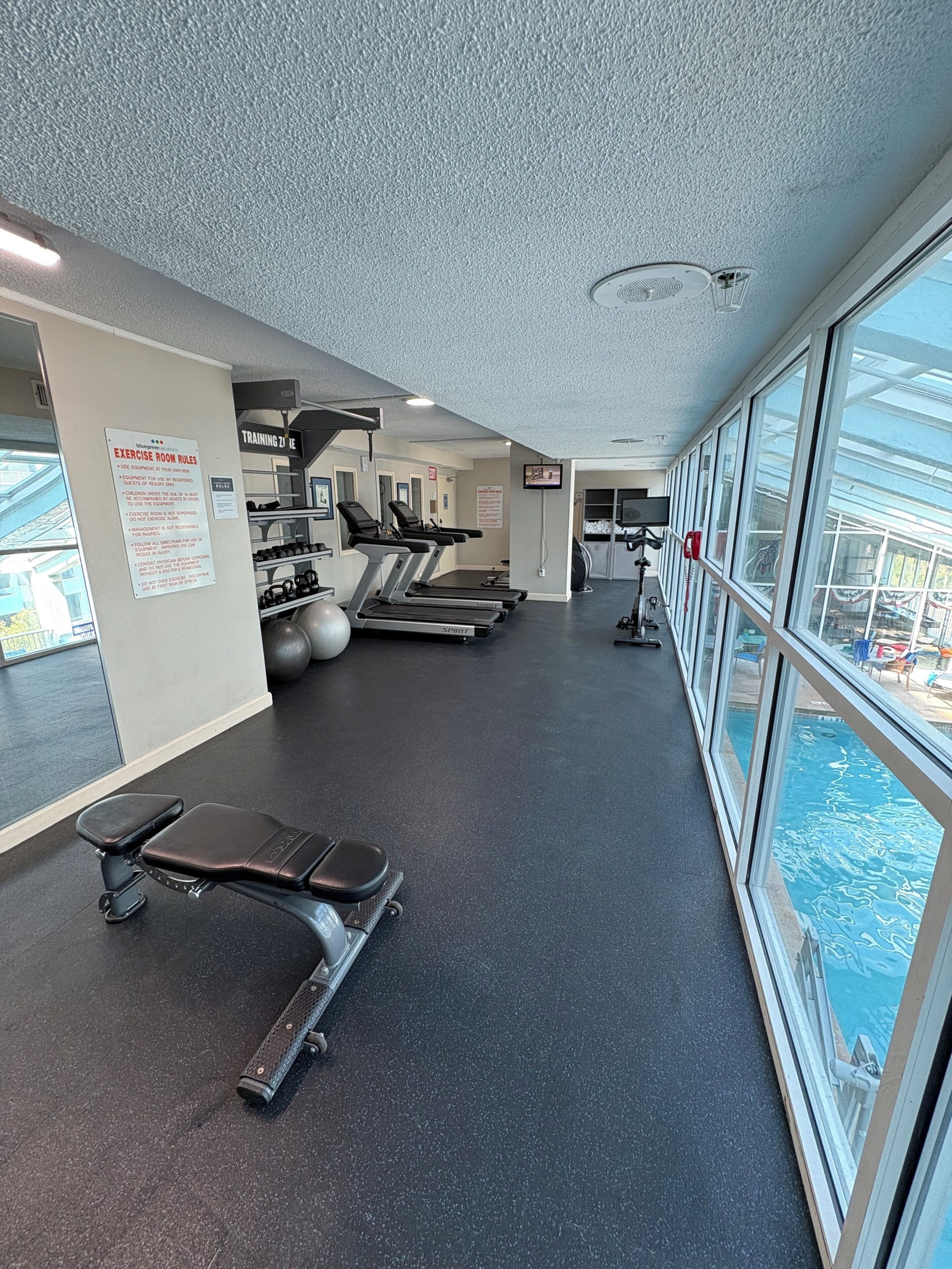 Une salle de sport équipée de tapis de course, d’un banc et d’une piscine, le tout visible à travers de grandes fenêtres.