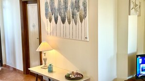 Un couloir avec une porte blanche, une œuvre d’art encadrée représentant des feuilles stylisées, une table avec une lampe et des objets décoratifs, et un téléviseur posé sur un meuble.