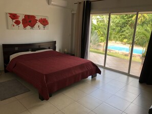 Une chambre à coucher avec un lit, un tableau représentant des fleurs rouges, une fenêtre donnant sur une piscine et sur de la verdure.