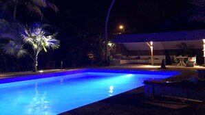 Ein Schwimmbecken bei Nacht mit einer Stein-Terrasse, Palmen und einer überdachten Sitzgruppe mit Tischen und Stühlen.
