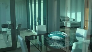 Un comedor moderno con una mesa y sillas de vidrio, un sofá, y vistas a un balcón a través de puertas de vidrio.