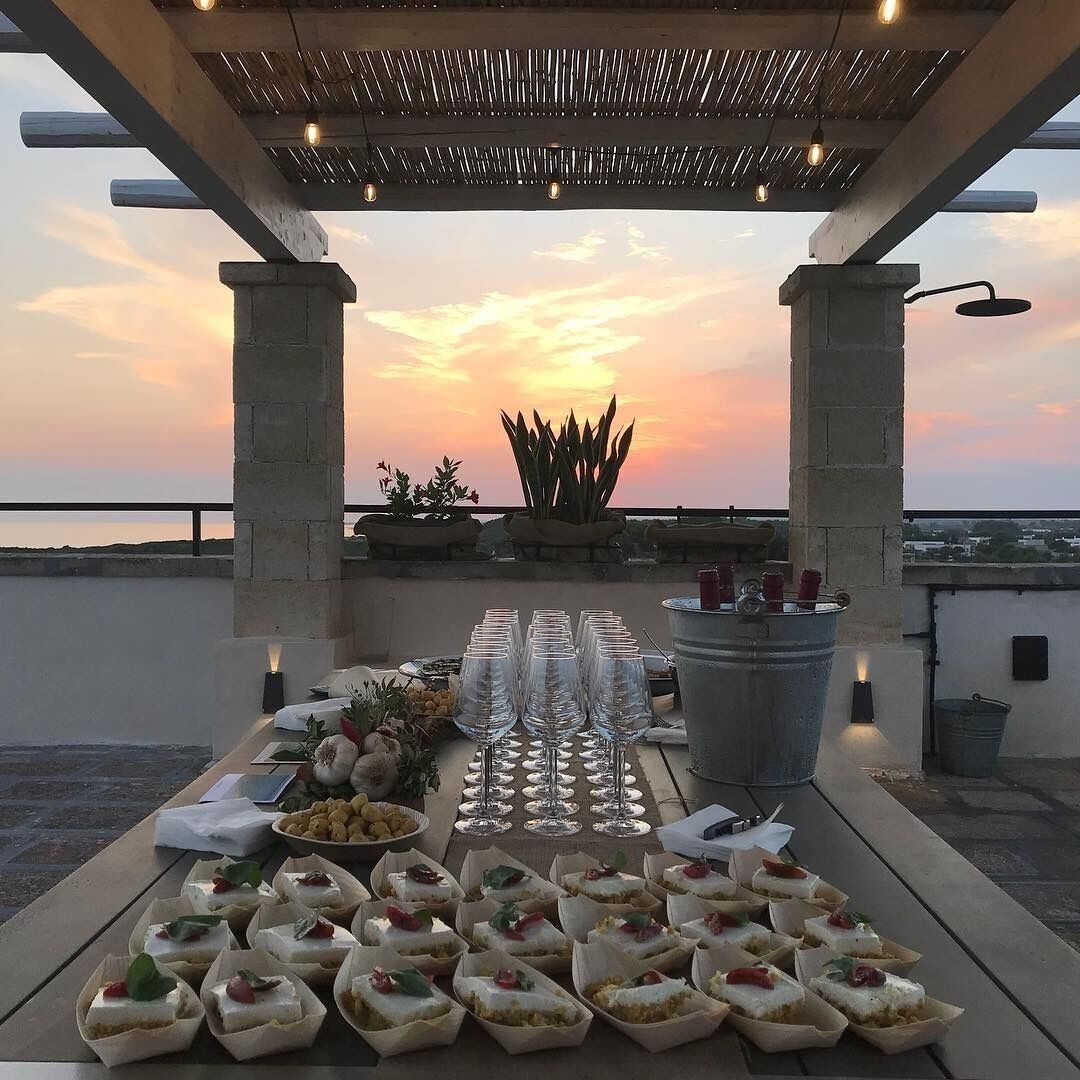 A rooftop dining setup with a sunset view, featuring a table with food, glasses, and a potted plant.