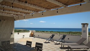 An outdoor patio with a table and chairs, a large vase, and lounge chairs facing a scenic ocean view.