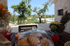 Une table avec des aliments pour le petit-déjeuner, un panier de fruits et une vue sur une piscine et des arbres.