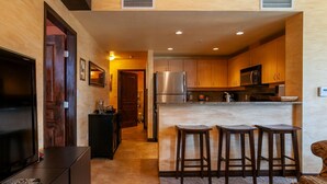Une cuisine compacte avec des meubles en bois, un réfrigérateur en acier inoxydable et un four à micro-ondes. Il y a deux tabourets au comptoir et un téléviseur sur le côté gauche.