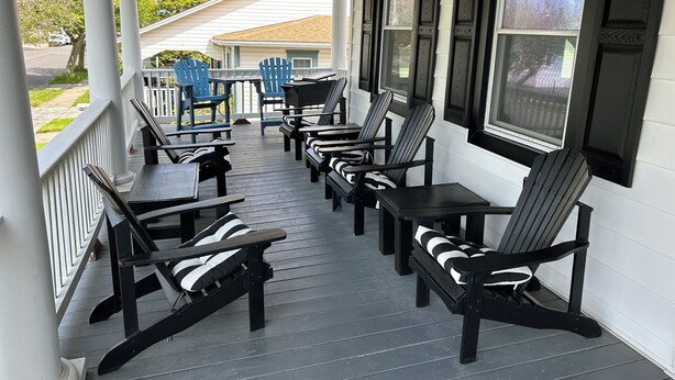 A covered porch with black and white Adirondack chairs and tables, white railings, and large windows with dark shutters.