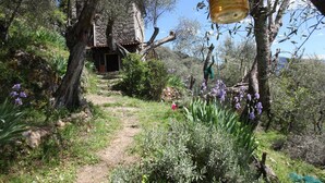 Un edificio rustico in pietra con un sentiero sterrato che vi conduce, circondato da vegetazione lussureggiante e fiori viola.
