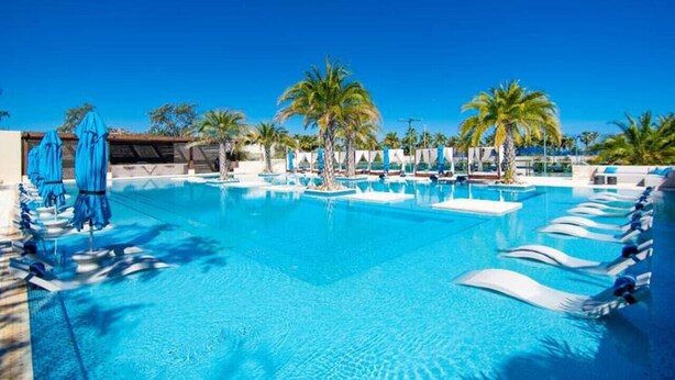 Una grande piscina esterna con numerosi lettini e ombrelloni, circondata da palme.
