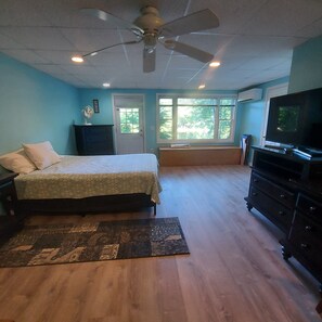 A bedroom with a bed, a dresser, a television, a ceiling fan, and a window with a view of greenery.