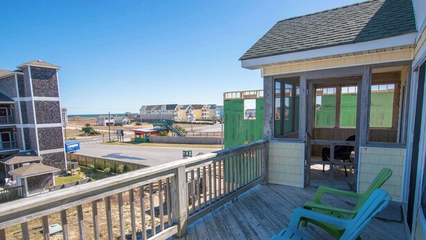 A wooden deck with two green chairs, a covered porch, and a view of the beach and other buildings. - 6 bedrooms, elevator,  3 minute walk to the beach. (Kill Devil Hills)