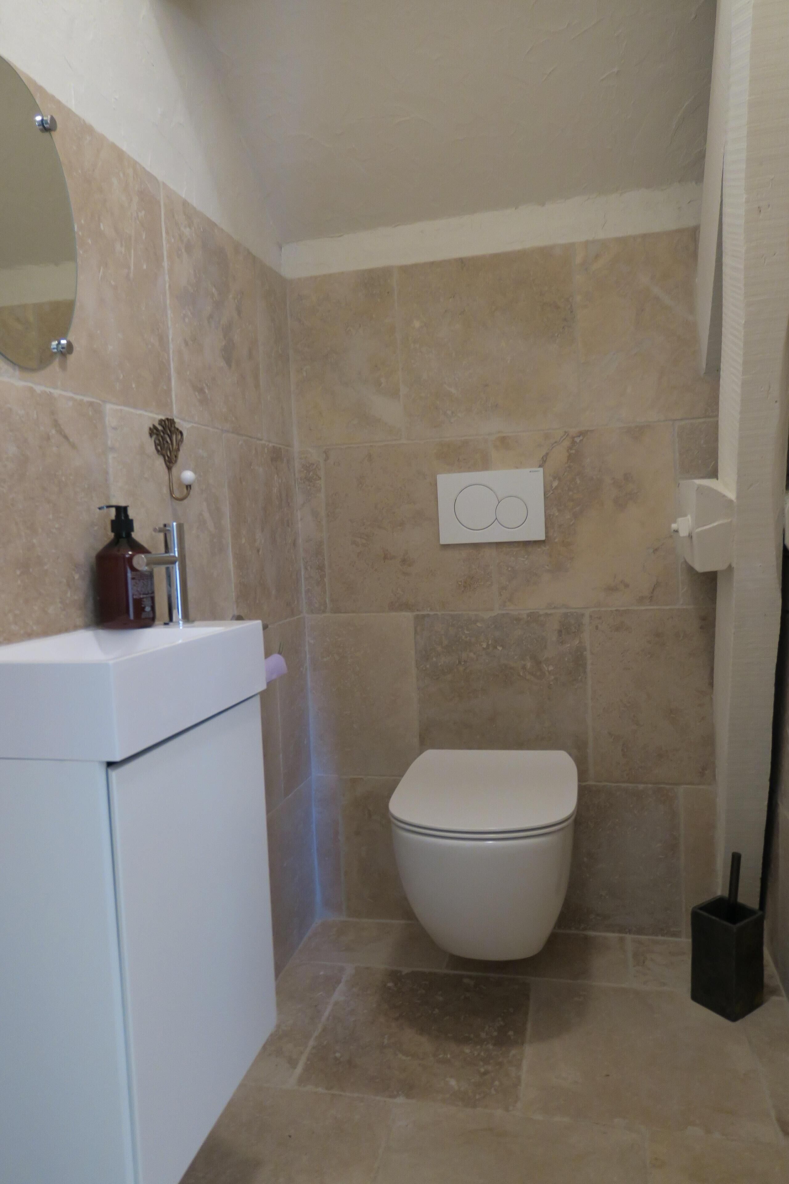 Un baño con baldosas color beige, un inodoro blanco, un lavamanos con dispensador de jabón y un espejo.