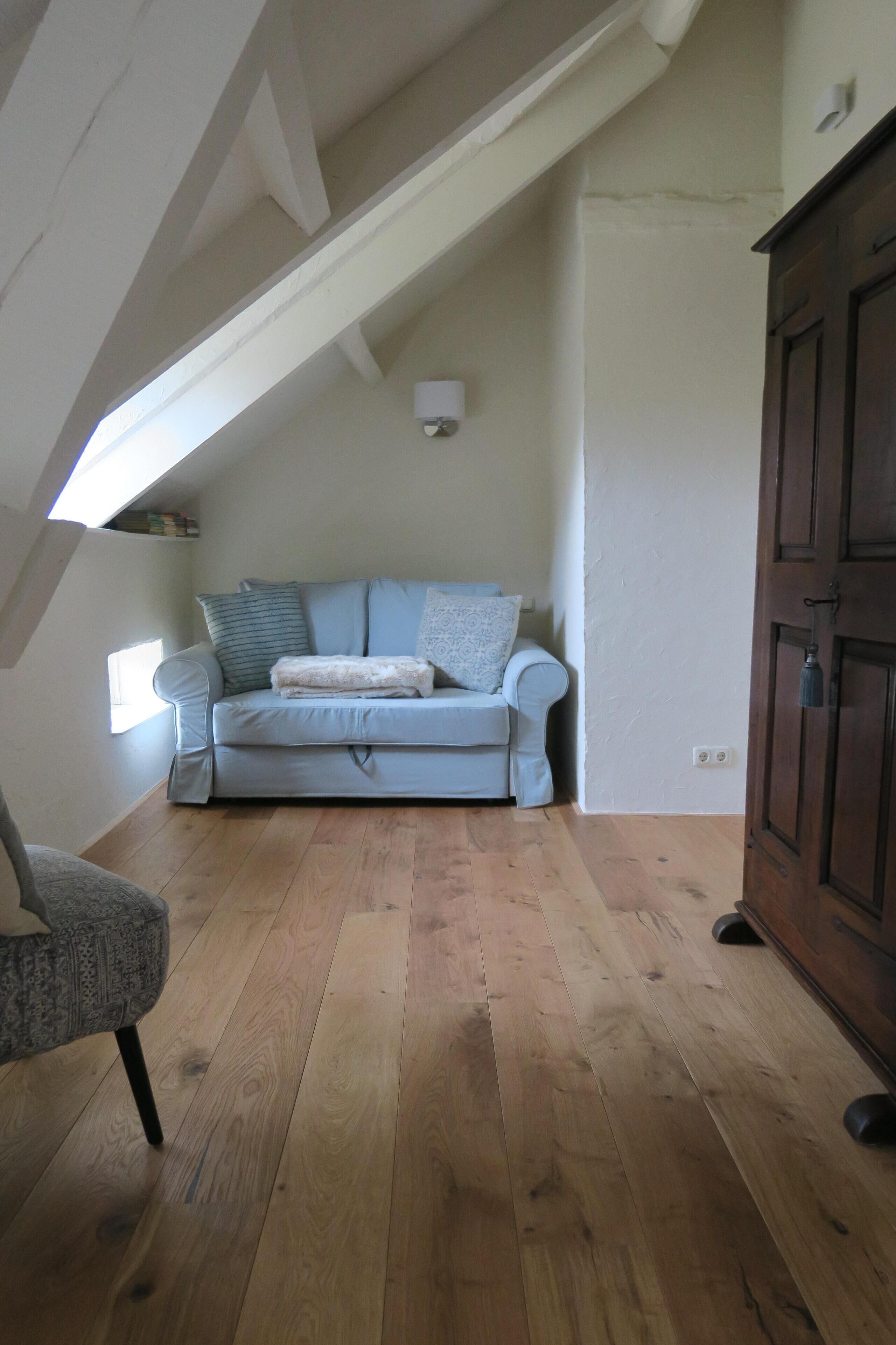 A loft-style room with a sloped ceiling, a light blue sofa with patterned cushions, a wooden floor, and a dark wooden cabinet.