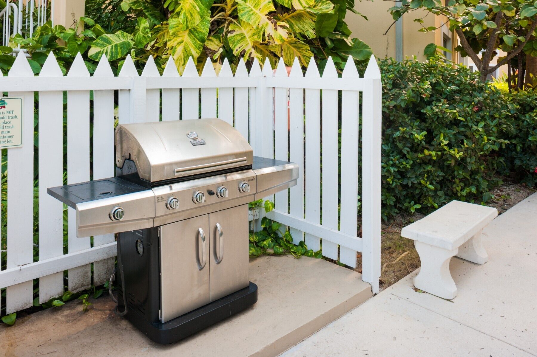A portable stainless steel grill with four burners and a side burner, placed on a concrete patio next to a white picket fence.