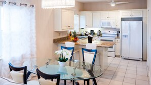 A modern kitchen with white cabinets, a stainless steel refrigerator, and a dining area with a glass table and blue chairs. - Regal Beach Club #231 (Seven Mile Beach)