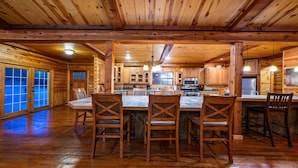 A wooden cabin interior with a central dining table, four chairs, wooden cabinets, a kitchen area with appliances, and large windows with blue-tinted glass.