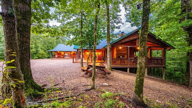 A cabin with a stone fireplace, wooden deck, and a gravel driveway surrounded by trees.