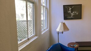 Een kamer met een groot raam, een blauwe stoel, een vloerlamp en een lijst met een foto aan de muur.