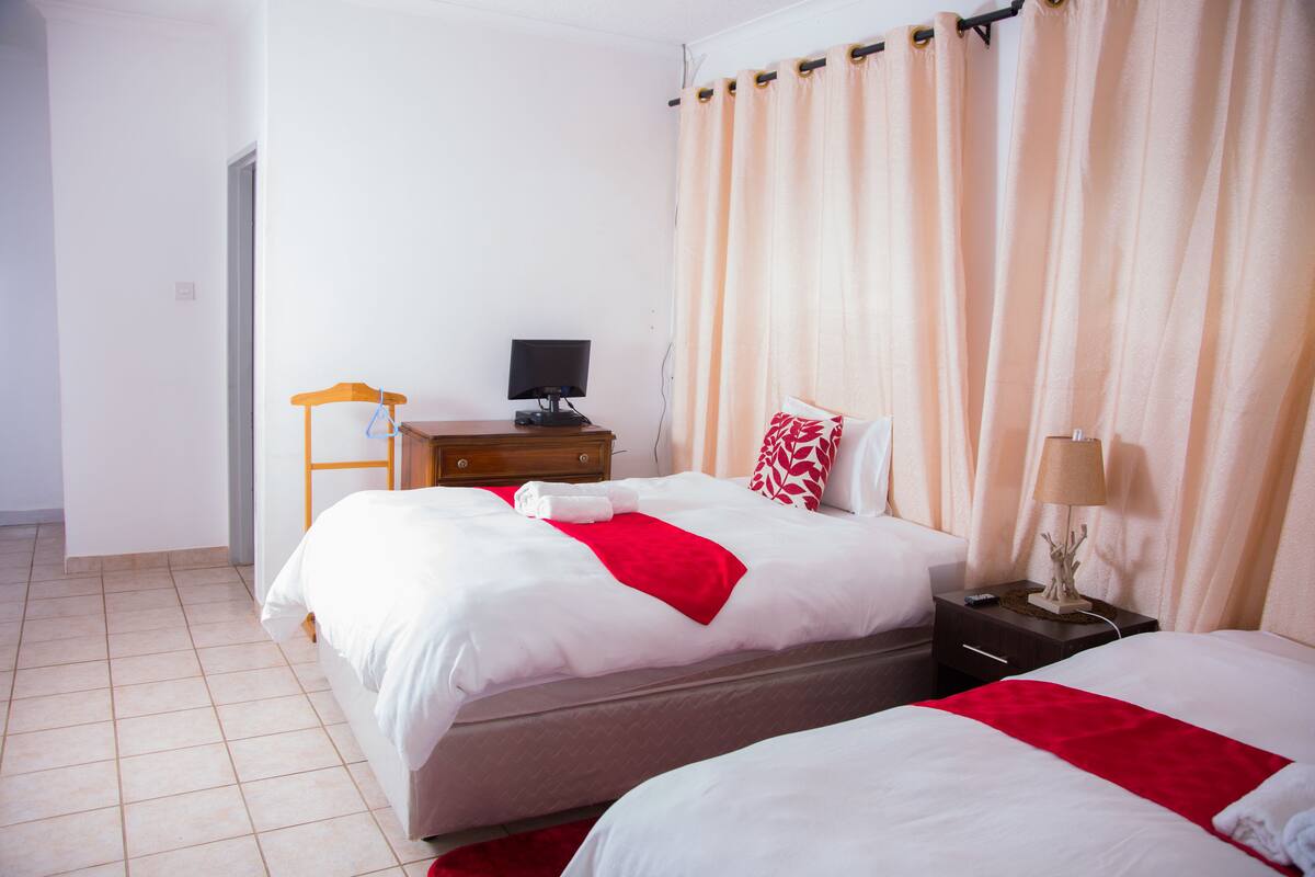 A hotel room with two beds, a desk with a computer, a lamp, and a window with curtains.