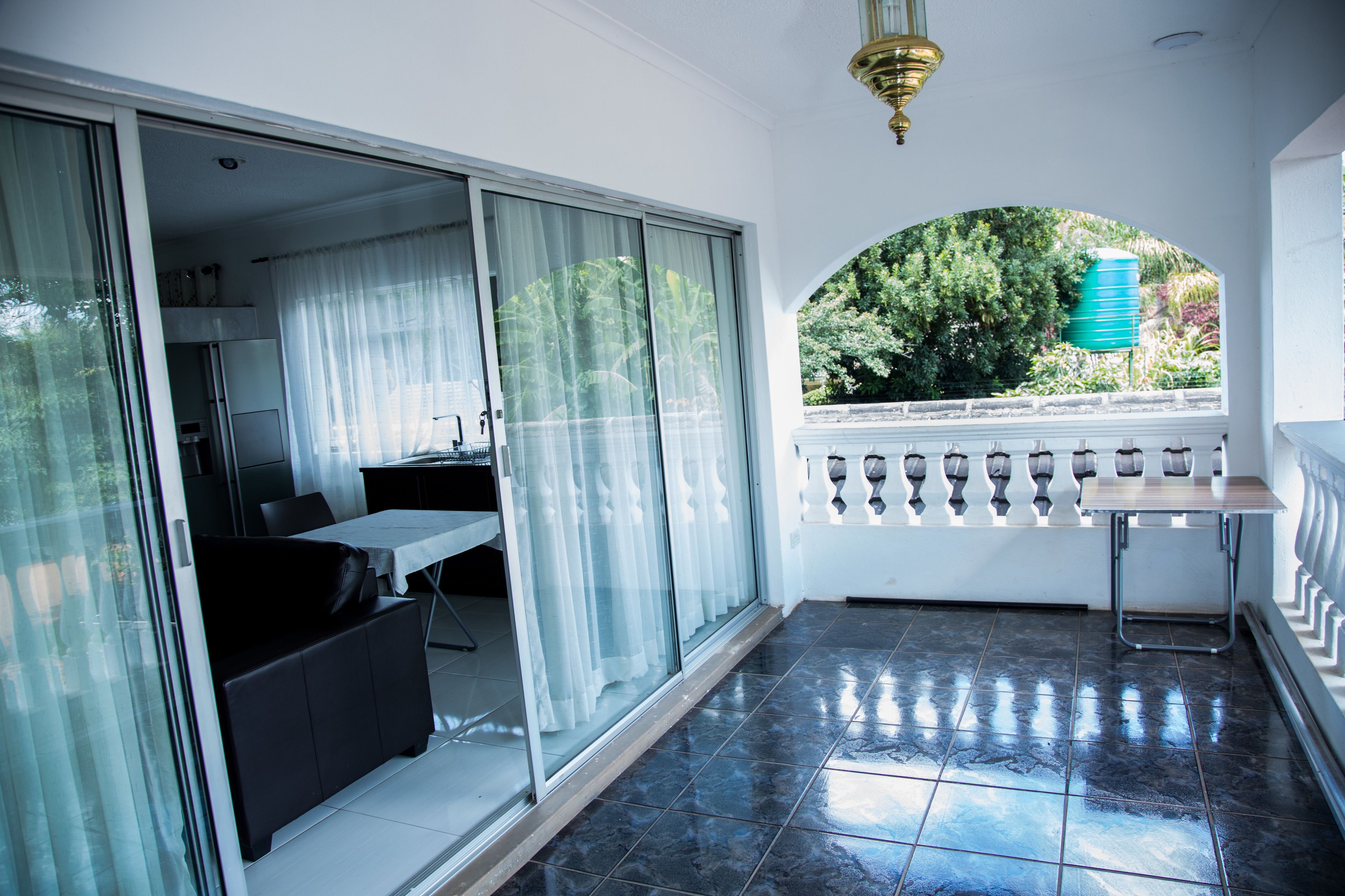 A balcony with a table and chairs, a sliding glass door leading to a room with a bed and a desk, and a view of greenery outside.