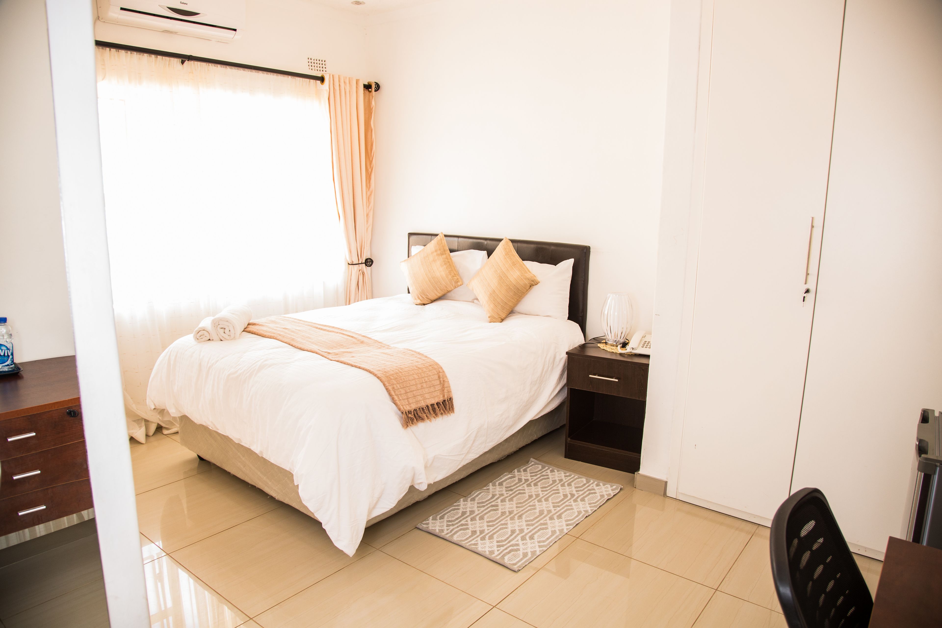 A neatly made bed with pillows, a bedside table with a lamp, and a patterned rug on a tiled floor.