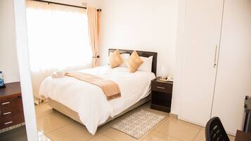 A neatly made bed with pillows, a bedside table with a lamp, and a patterned rug on a tiled floor.