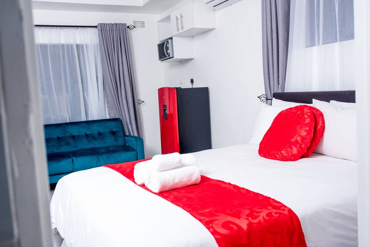 A hotel room with a bed, a red and white bedspread, a blue chair, a wardrobe, and a small TV on a shelf.