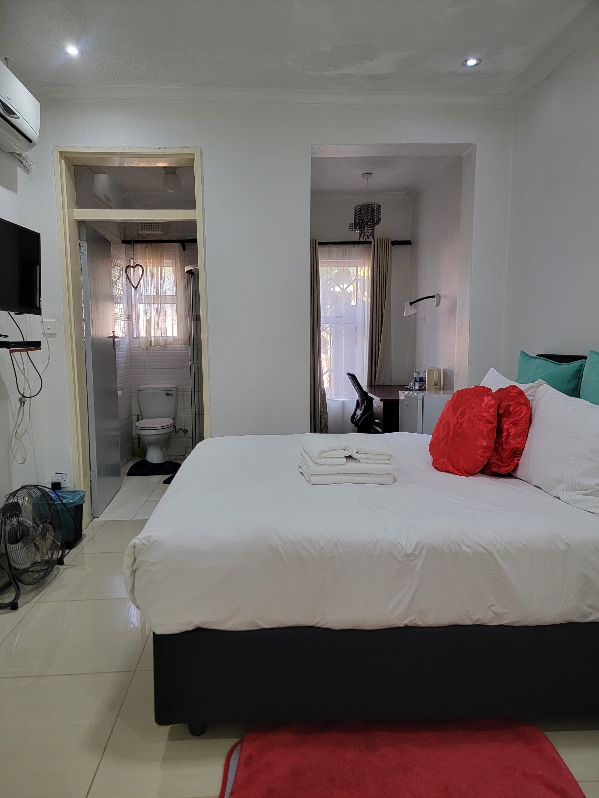 Habitación de hotel con cama, un banco rojo, televisor, ventilador y baño visible a través de una puerta abierta.