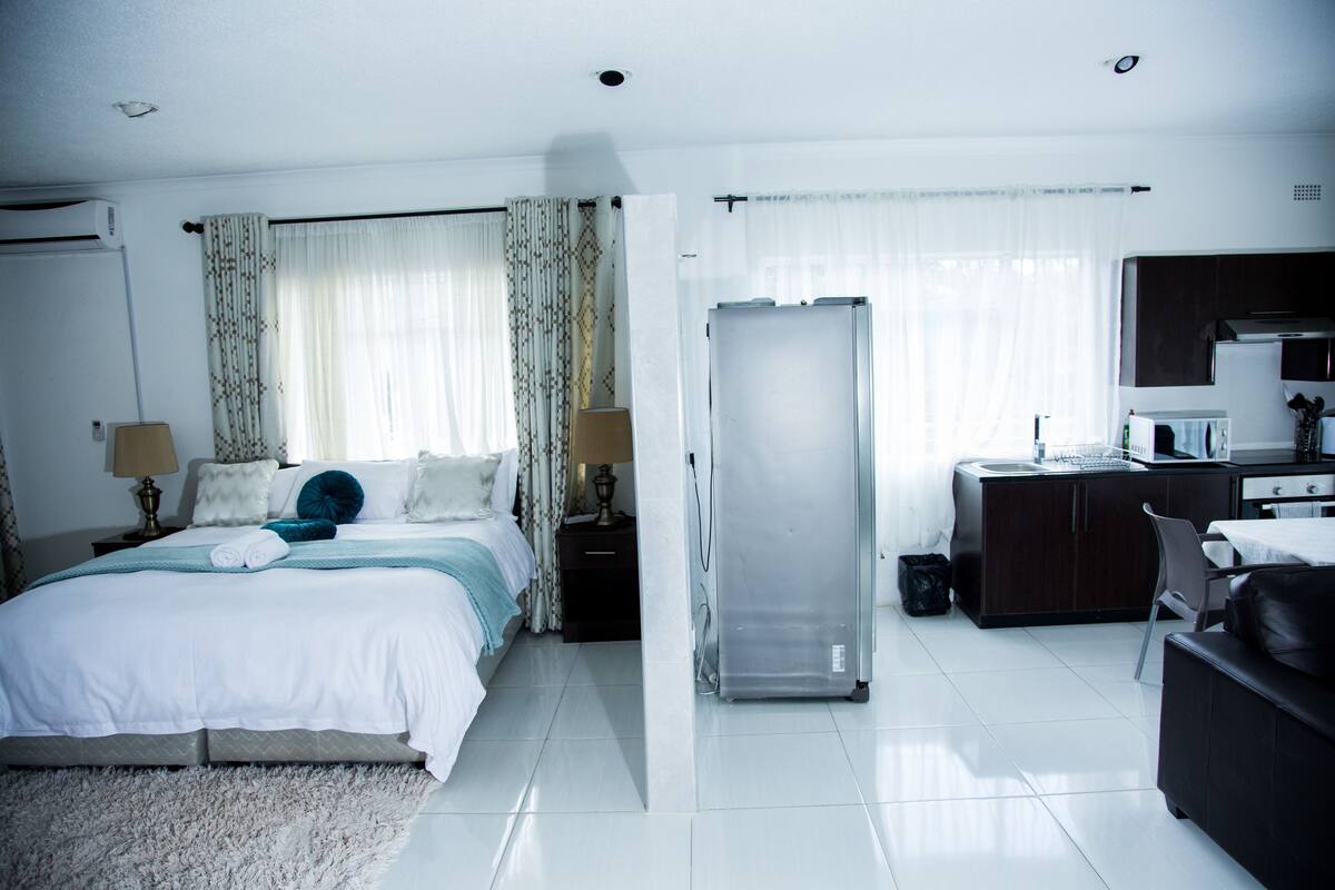A modern hotel room with a large bed, a separate bathroom, a kitchenette, and a view of the city.