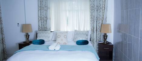 A neatly made bed with a blue blanket, two bedside lamps, and a window with sheer curtains.