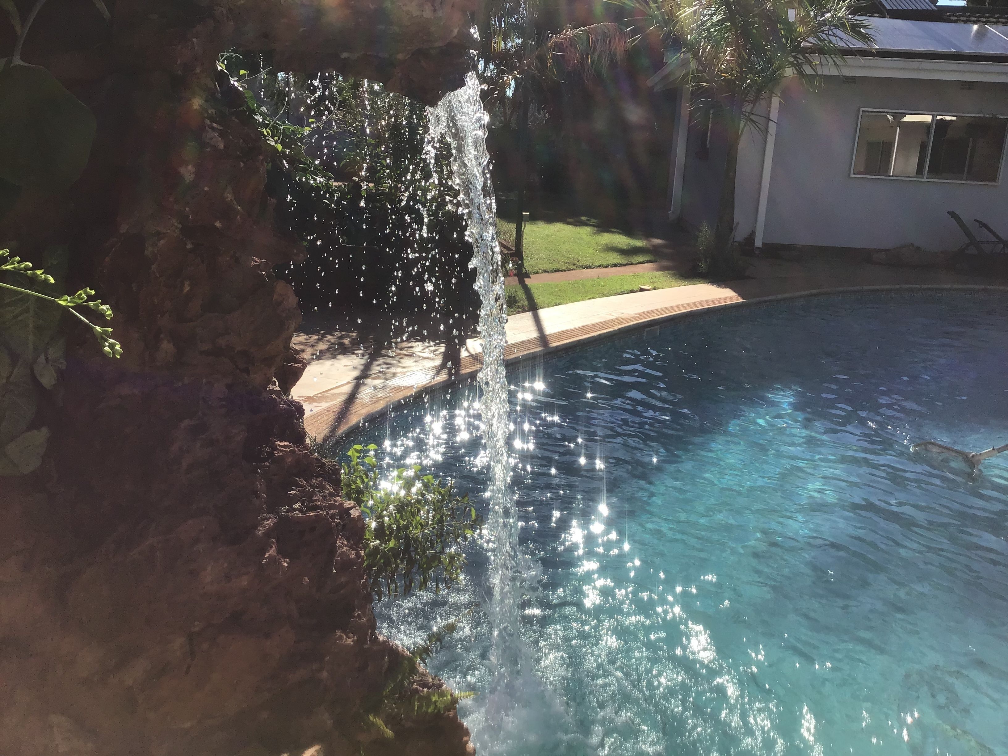 A swimming pool with a waterfall feature, surrounded by a grassy area and a white building.