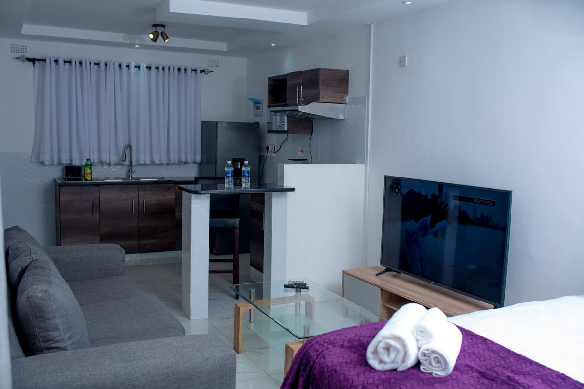 A modern hotel room with a grey sofa, a glass coffee table, a TV, a bed with a purple blanket, and a kitchenette area.